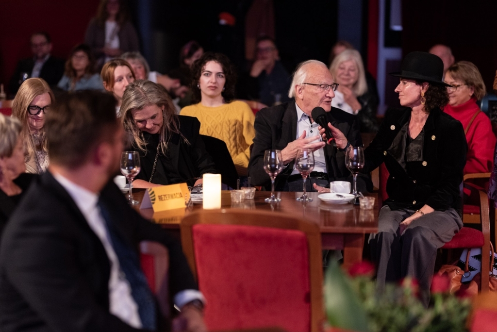 Przy stoliku siedzą kobiety i mężczyźni, jeden mężczyzna odpowiada do mikrofonu