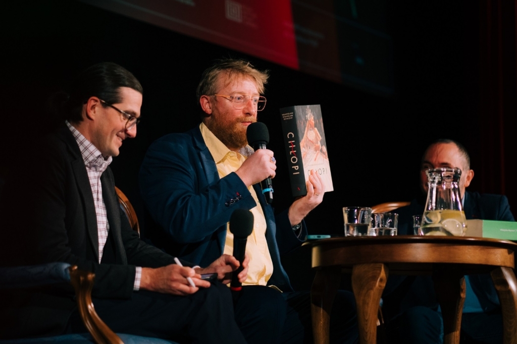 Na scenie mężczyźnie siedzący przy stole, prezentujący książkę Chłopi