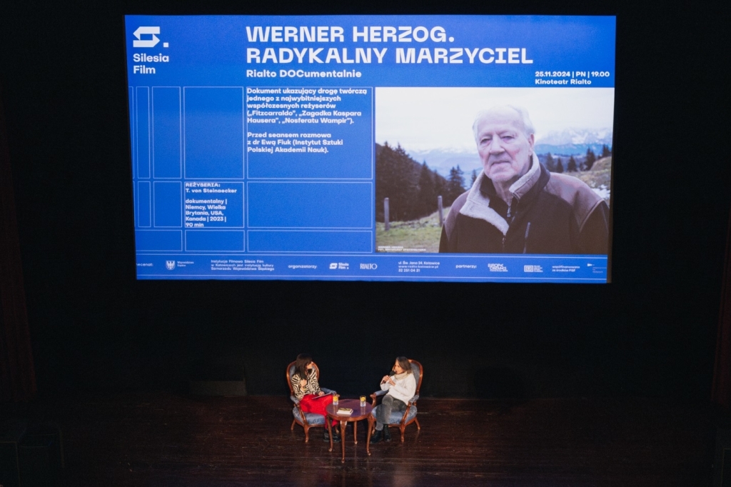 Na scenie dwie kobiety siedzące na krzesłach na ekranie wyświetlony napis Werner Herzog radykalny marzyciel